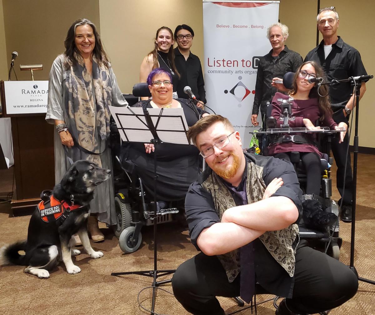 Eight people posing in front of a Listen to Dis' banner and a podium, smiling at the camera, also joined by a service dog in training. Two of the people in the photo are seated in wheel chairs and have microphones; one person in the front is squatting with their arms crossed; the rest are standing.