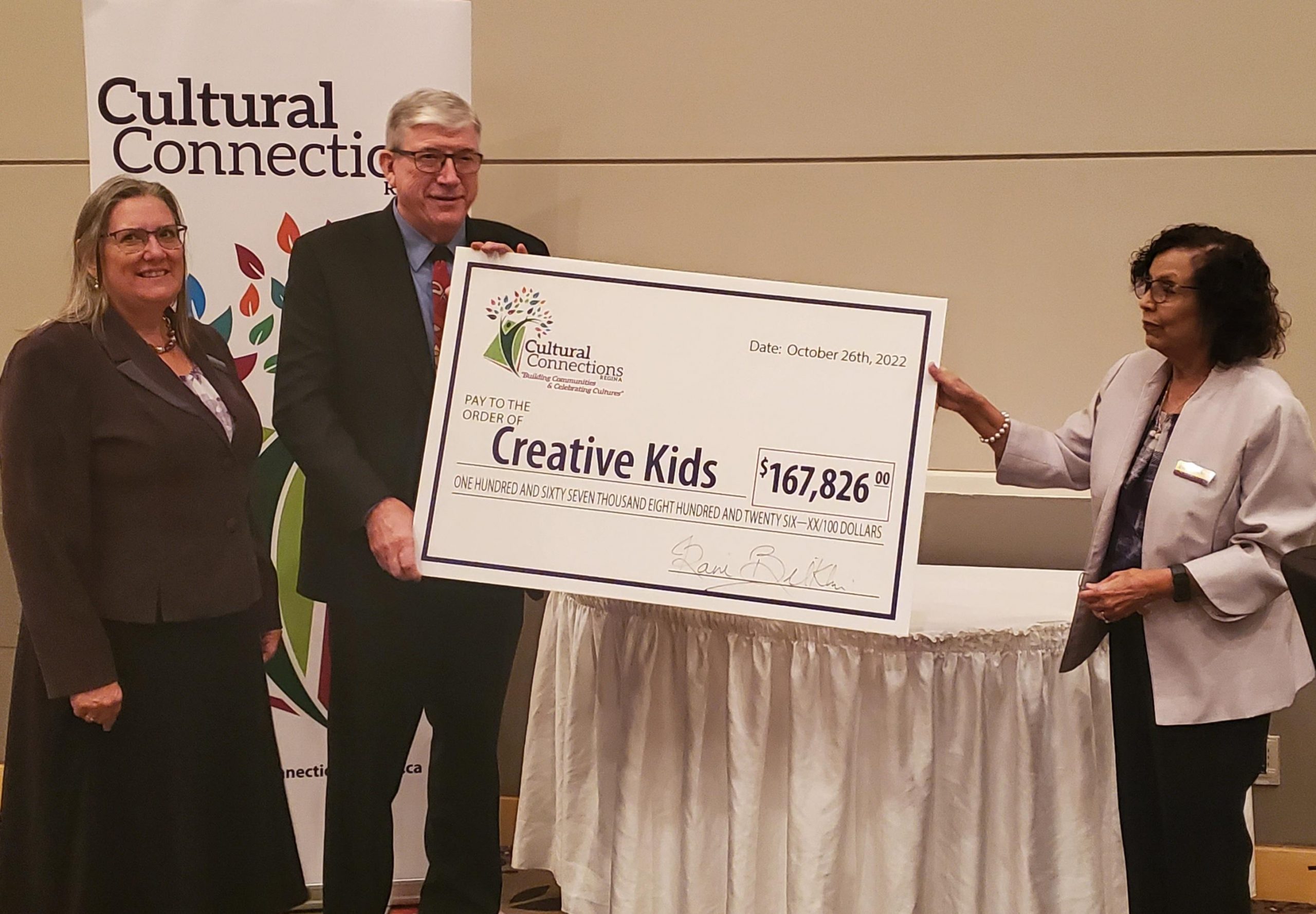 Three people posing in front of a Cultural Connections banner. Two of them are holding a big cheque with the Cultural Connections logo, which reads "Pay to the order of Creative Kids," for the amount of $167,826.