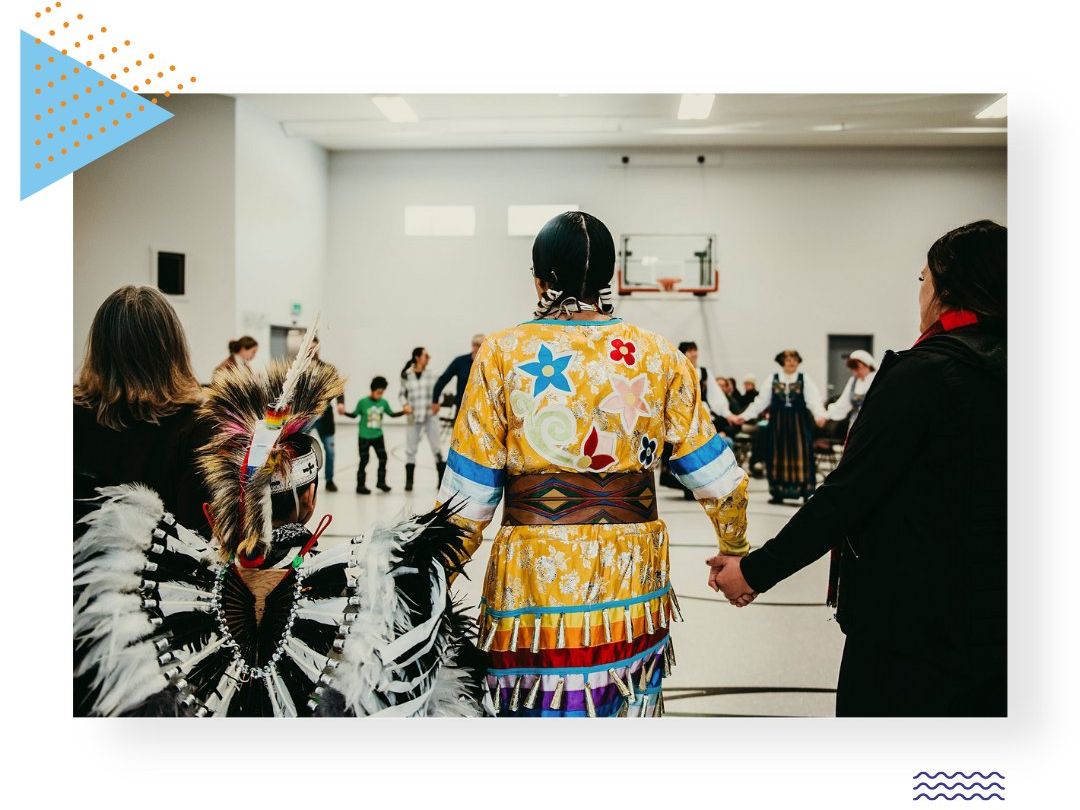 A photo of a powwow. The two people closest to the camera are a kid and an adult wearing traditional regalia.