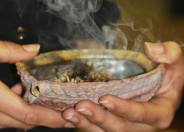 A photo of someone smudging, holding a shell that contains smoldering plants.
