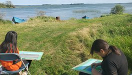 Two young people from English River First Nation’s sitting on chairs in the grass, in front of water, taking part in Summer Land-Based Learning.