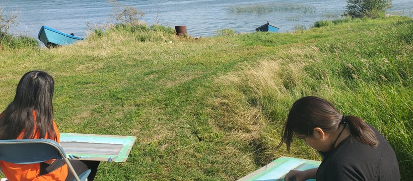 Two young people from English River First Nation’s sitting on chairs in the grass, in front of water, taking part in Summer Land-Based Learning.