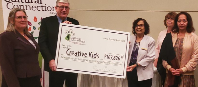 Three people posing in front of a Cultural Connections banner. Two of them are holding a big cheque with the Cultural Connections logo, which reads "Pay to the order of Creative Kids," for the amount of $167,826.