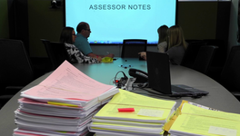 A photo of people in a conference room. A Power Point presentation in the back reads: "Assessor Notes."
