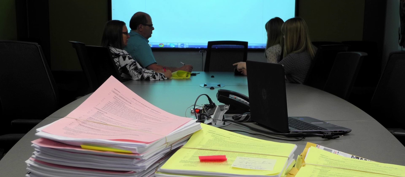 A photo of people in a conference room. A Power Point presentation in the back reads: "Assessor Notes."