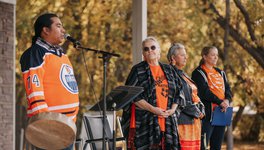 A group of four people wearing orange clothing. On the left, Lindsay Littlechief talks on a mic about the significance of the Honour song. Elder Angie McArthur-Delorme is second from the right.