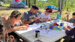 Participants and Knowledge Keepers from this year’s OUT on the Land camp sitting around table beading. The background shows trees and camping tents.