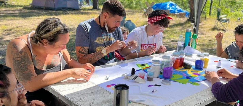 Participants and Knowledge Keepers from this year’s OUT on the Land camp sitting around table beading. The background shows trees and camping tents.