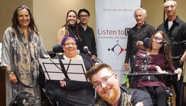 Eight people posing in front of a Listen to Dis' banner, smiling at the camera, also joined by a service dog in training. Two of the people in the photo are seated in wheel chairs and have microphones; one person in squatting; the rest are standing.