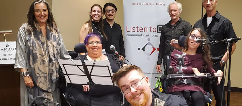 Eight people posing in front of a Listen to Dis' banner, smiling at the camera, also joined by a service dog in training. Two of the people in the photo are seated in wheel chairs and have microphones; one person in squatting; the rest are standing.