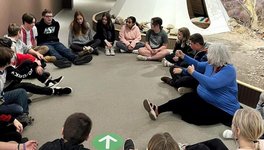 A photo of, Barb Frazer, Knowledge Holder, and a group of kids, all sitting on the floor in a circle. In the background there is a tipi.