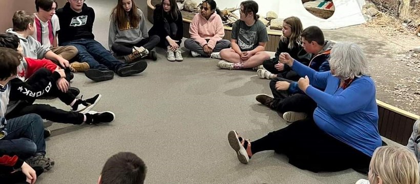 A photo of, Barb Frazer, Knowledge Holder, and a group of kids, all sitting on the floor in a circle. In the background there is a tipi.