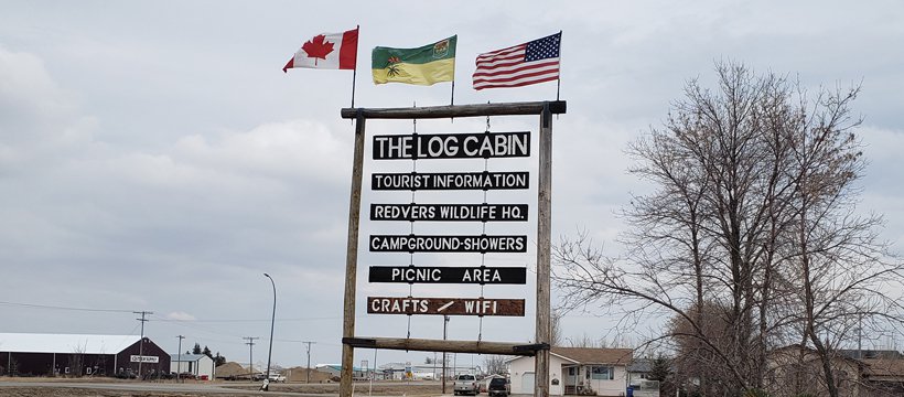 A photo of a sign that reads "The Log Cabin" in Redvers that offers tourist information.