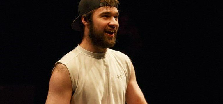 A photo of John Loeppky performing in a play. Loeppky is barefoot and wearing athletic wear, sitting on a wheelchair.