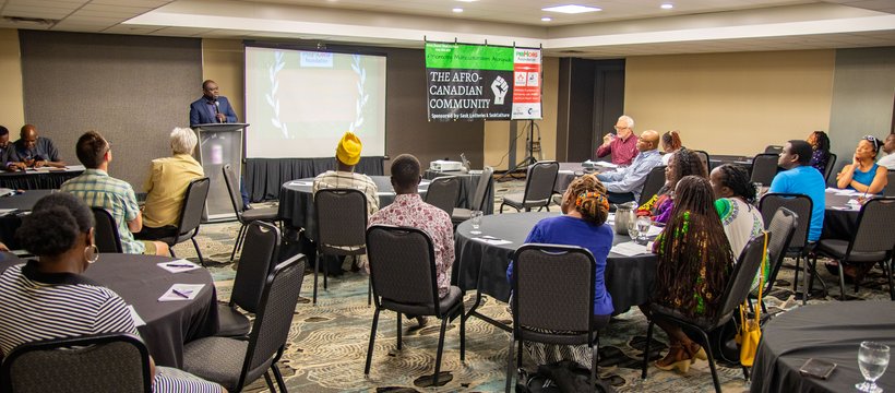 A photo of PRIMORG's first event. it shows people gathered in an events room, looking at a speaker at the podium. Next to the speaker there is a poster that reads: The Afro-Canadian Community.