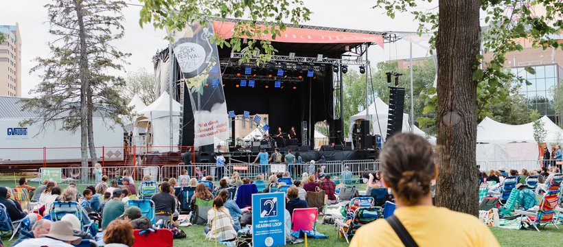 A photo of the crowd gathered on the lawn in front of a big festival stage. A light blue sign in the middle of the lawn advertises a hearing loop.