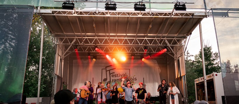 A photo of a group of performers on a festival mainstage. The performers are holding instruments and singing into microphones. In the back there is a big sign that reads "Festival Fransaskois."