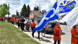 A crowd of people wearing orange shirts, marching with posters, drums and flags, including the Métis flag.