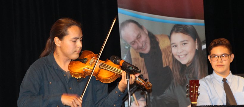 A musician plays a fiddle in front of a microphone while a panelist watches.