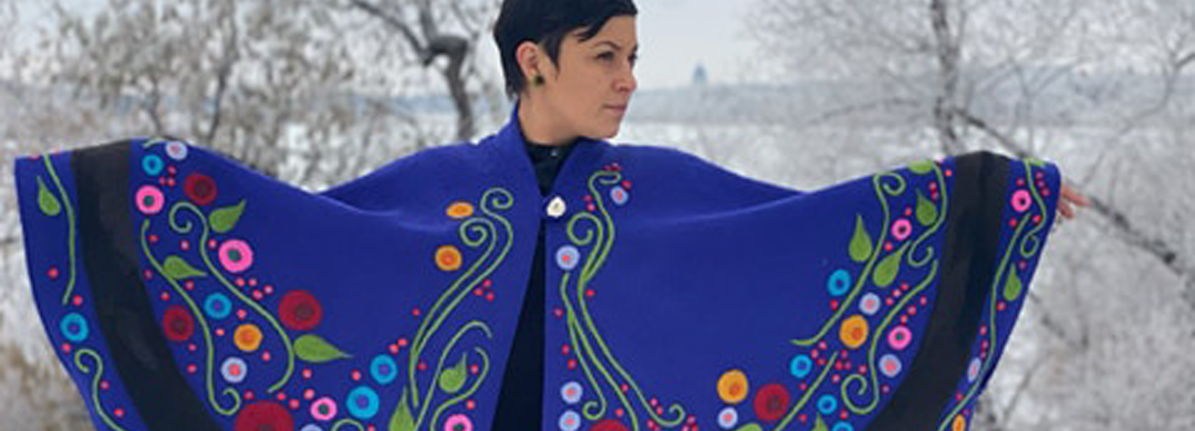 A person stands in a snowy field displaying an intricate blue and multicoloured cloak on their body.