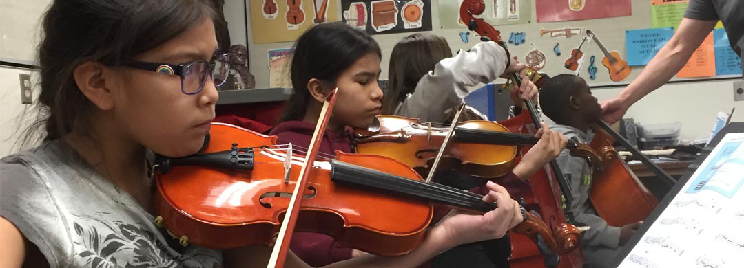 A collection of youth play violins in a classroom.