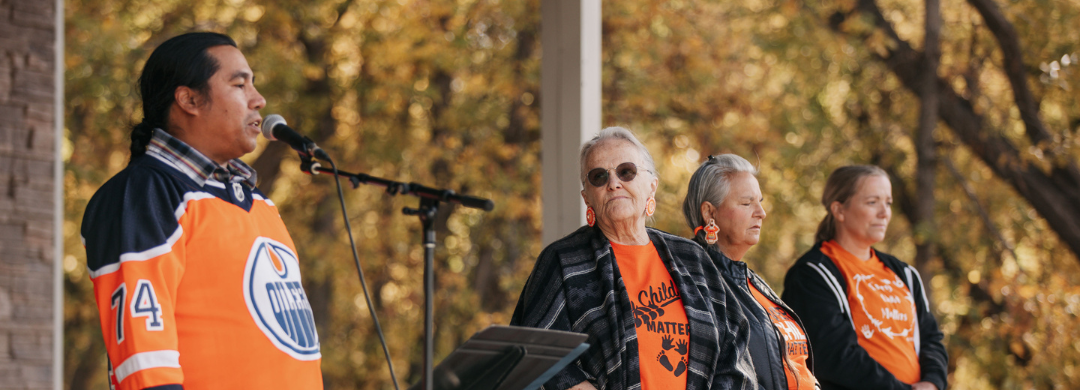 A group of four people wearing orange clothing. On the left, Lindsay Littlechief talks on a mic about the significance of the Honour song. Elder Angie McArthur-Delorme is second from the right.