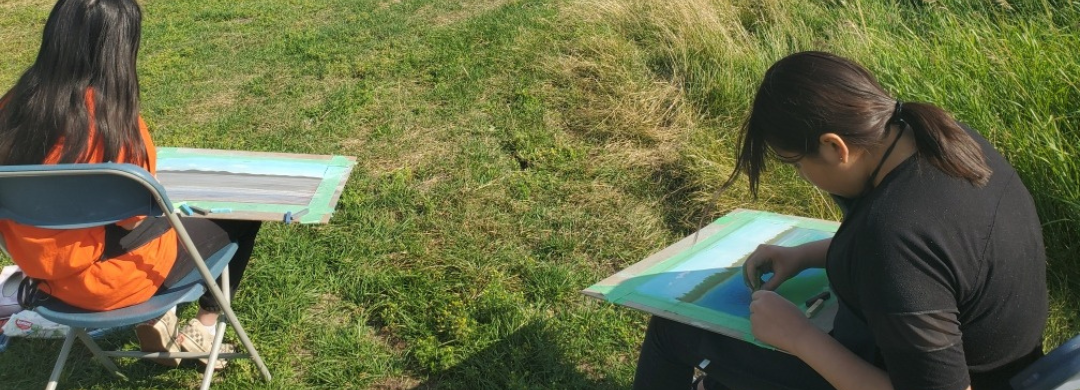 A photo of two kids sitting outdoors facing the water, drawing the landscape in front of them.
