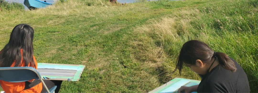 A photo of two kids sitting outdoors facing the water, drawing the landscape in front of them.