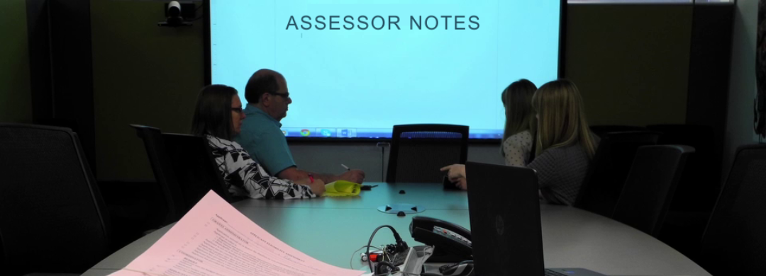 A photo of people in a conference room. A Power Point presentation in the back reads: "Assessor Notes."