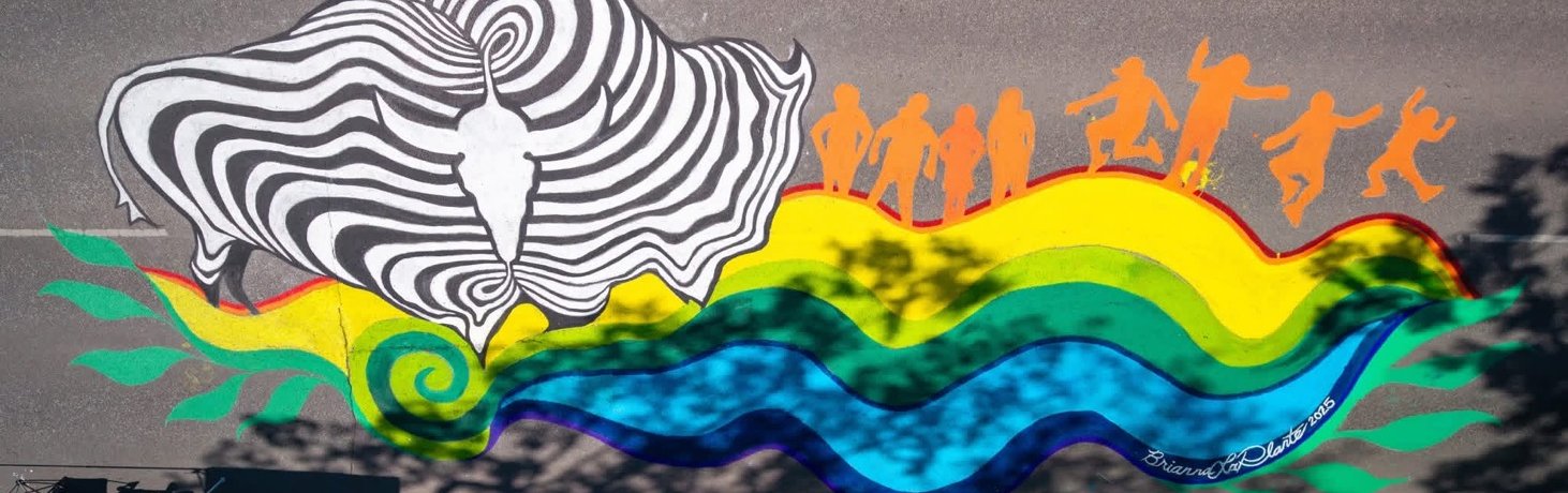 A colorful street mural features a large black-and-white abstract design resembling a buffalo head on the left, with swirling lines. To the right, orange silhouettes of dancing and jumping figures are positioned above a flowing rainbow wave pattern