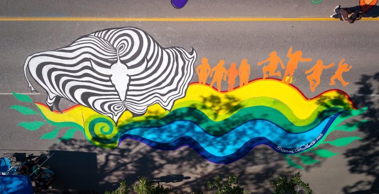 A colorful street mural features a large black-and-white abstract design resembling a buffalo head on the left, with swirling lines. To the right, orange silhouettes of dancing and jumping figures are positioned above a flowing rainbow wave pattern