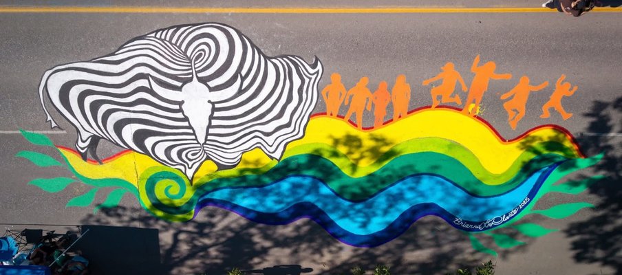 A colorful street mural features a large black-and-white abstract design resembling a buffalo head on the left, with swirling lines. To the right, orange silhouettes of dancing and jumping figures are positioned above a flowing rainbow wave pattern in gre