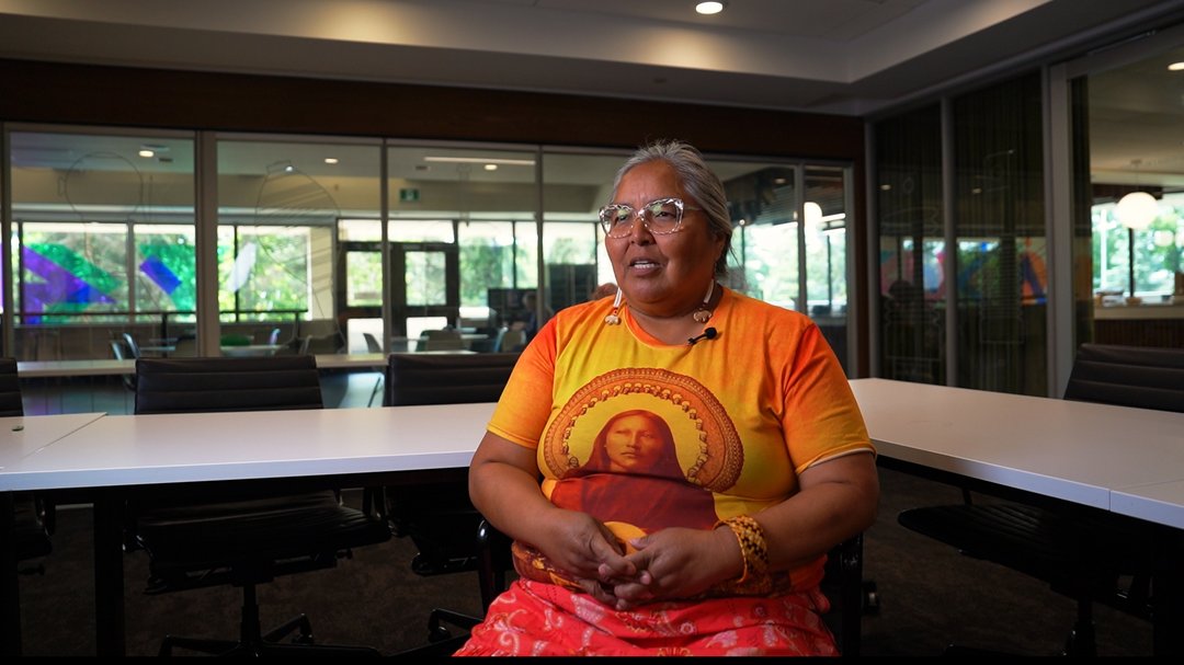 A person sits at a white conference table in a modern meeting room with large windows, wearing a bright orange shirt with a circular design and a red patterned skirt, with greenery visible outside.