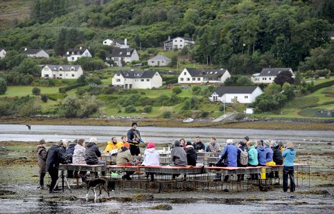 A group of people gather around tables set on a tidal shoreline while a guide speaks at the center. Across the water, a small village and forested hillside create a scenic backdrop.