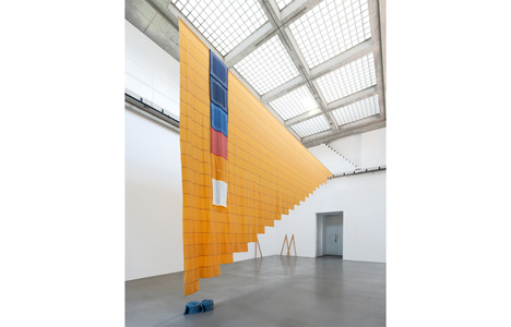 A large textile installation hangs from the ceiling of a bright gallery with a grid-patterned skylight. The suspended artwork features layered orange fabric with blue, red, and white elements, cascading diagonally across the open space.