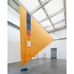 A large textile installation hangs from the ceiling of a bright gallery with a grid-patterned skylight. The suspended artwork features layered orange fabric with blue, red, and white elements, cascading diagonally across the open space.