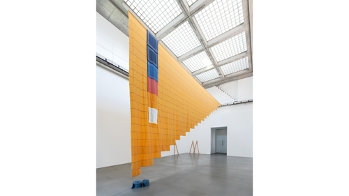 A large textile installation hangs from the ceiling of a bright gallery with a grid-patterned skylight. The suspended artwork features layered orange fabric with blue, red, and white elements, cascading diagonally across the open space.