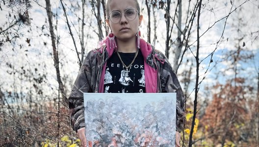 A photo of Kokum Bannock, also known as Janice Grayson, standing on a forest, holding up a painting.