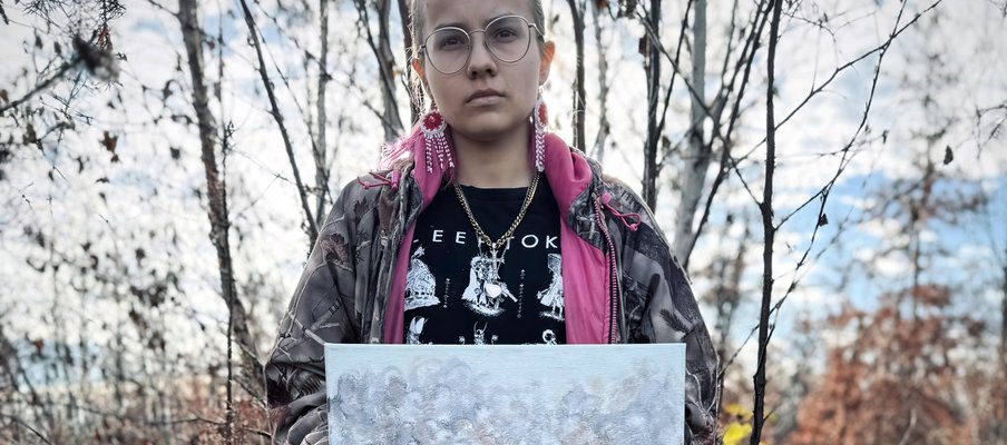 A photo of Kokum Bannock, also known as Janice Grayson, standing on a forest, holding up a painting.