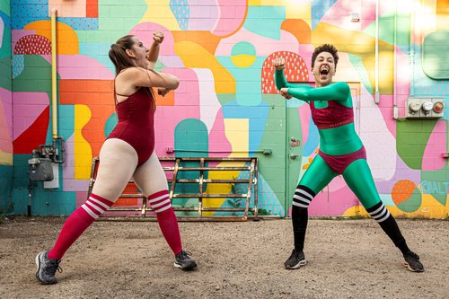 Two dancers move excitedly against a colourful mural on a building