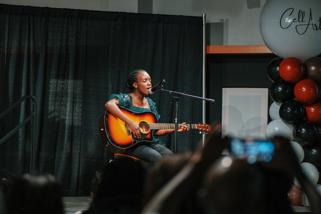 Person seated on stage playing an acoustic guitar in front of a microphone, with black curtain backdrop and balloon arrangement reading “Cell Art.”