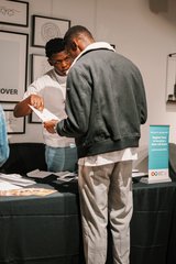 Two people stand at a table covered with papers and brochures, reviewing a document together.