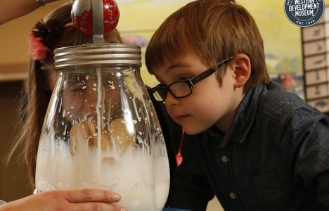 Two children watching a volunteer churn butter in a clear glass butter churn.