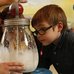 Two children watching a volunteer churn butter in a clear glass butter churn.
