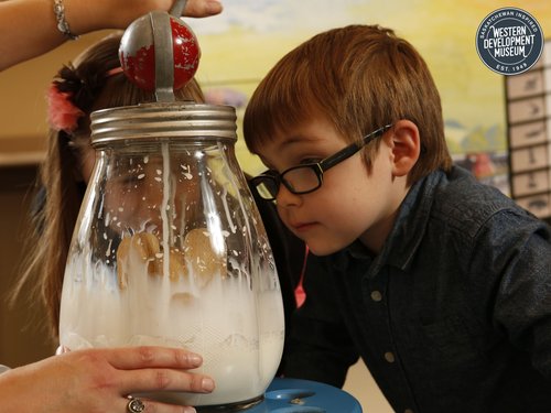 Two children watching a volunteer churn butter in a clear glass butter churn.