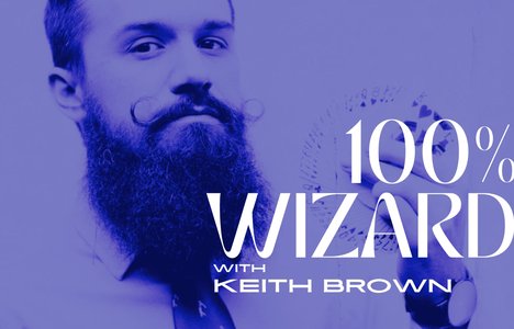Bearded Magician Keith Brown holds a fan of playing cards near his face. He's seen from the chest up. The entire image is in blue. There is white text overlayed on top that says 100% Wizard with Keith Brown