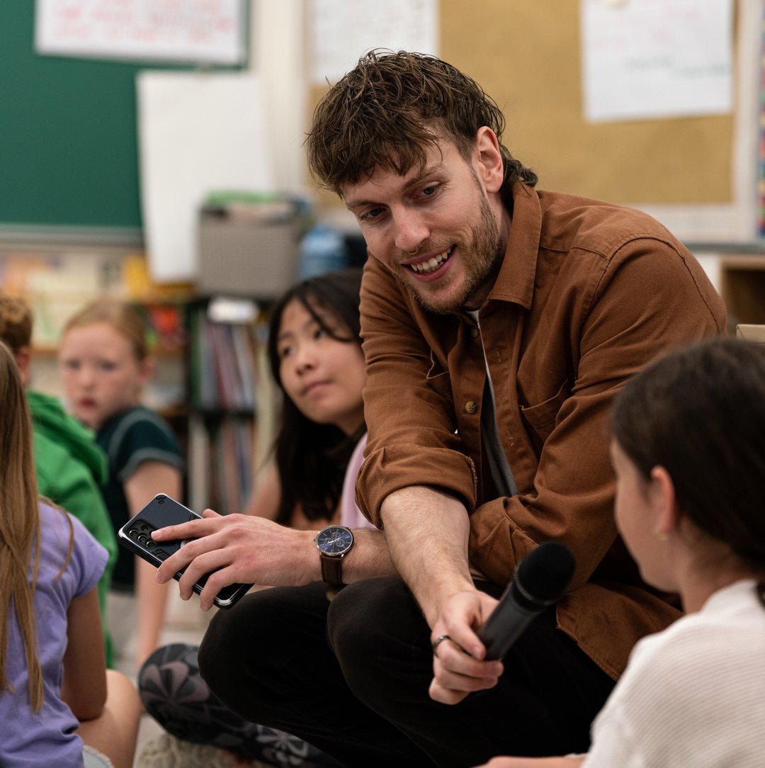 A person is seated in a classroom holding a microphone and a notebook, speaking with a child in the foreground. Other children are sitting nearby, and the background shows a chalkboard and bulletin boards.