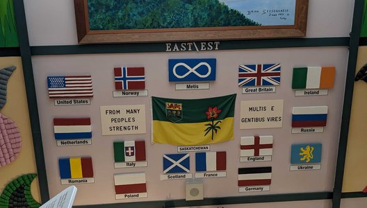 Exhibit wall featuring a framed painting of a bird flying over a winding river landscape at the top, with a display below showing various flags with languages to that the tours are offered in.
