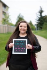 A person is standing outdoors on a paved pathway holding a sign with colorful text that reads “SLAVA UKRAINI! HEROYAM SLAVA.” The background shows grass, trees, a fence, and part of a building.