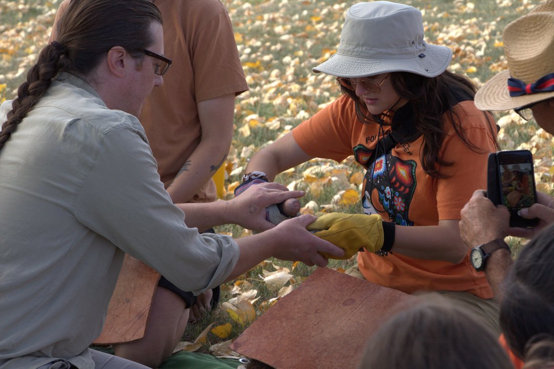 Two people kneeling on grass covered with fallen leaves, working together on breaking a rock to create flint arrowheads. One person wears an orange shirt and yellow gloves, holding an rock while another assists.